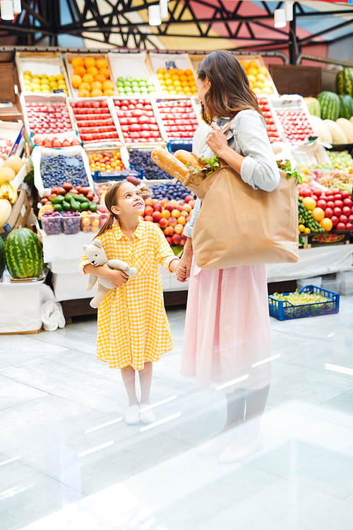 Young mother and pretty daughter standing in organic food store and holding hands while talking and buying fresh products together