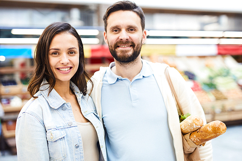 Happy positive young couple embracing and  while standing in food shop, smiling bearded man holding shopping bag on shoulder