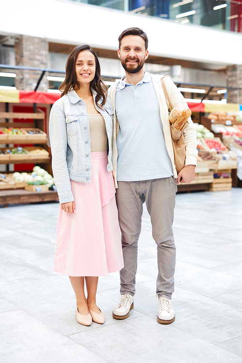 Cheerful beautiful young couple in casual clothing standing in farmers market and smiling at camera, they buying food together