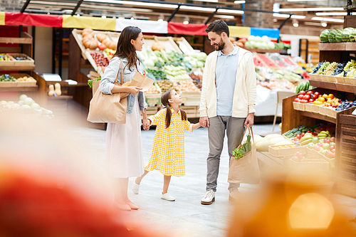 Happy beautiful young family in casual clothing holding hands and chatting together while purchasing food and walking over farmers market