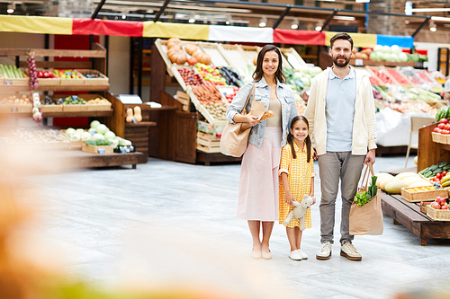 Positive beautiful young family in casual clothing standing in farmers market and holding shopping bags while 
