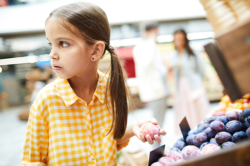 Worried girl with pony tails wearing checkered shirt standing at food shelves and holding plum while stealing it in food store