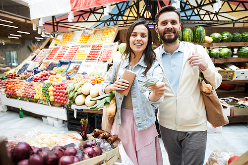 Positive beautiful young couple in casual clothing standing at farmers market and , while paying money for purchase