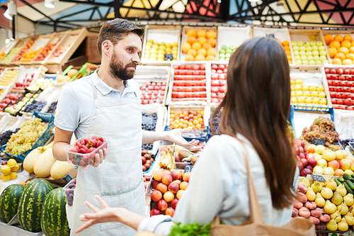 Confused misunderstanding young male grocer in apron holding container of strawberries shrugging shoulders while talking to customer in farmers market