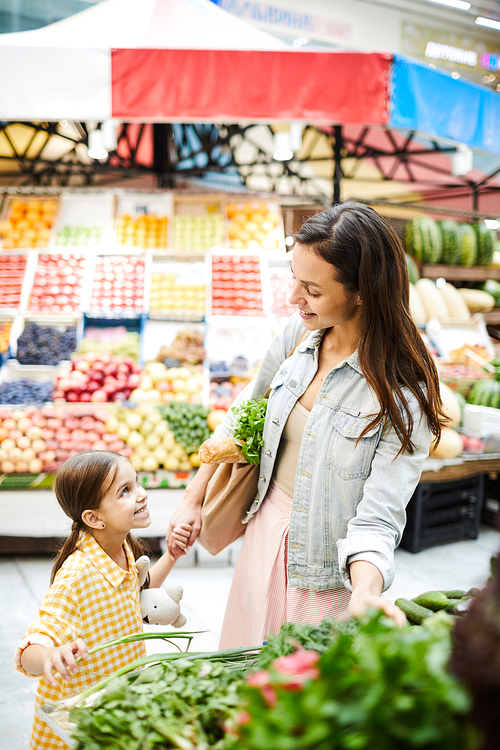 Smiling content attractive young mother in casual jacket standing at fresh food stall and choosing vegetables while talking to daughter in food store