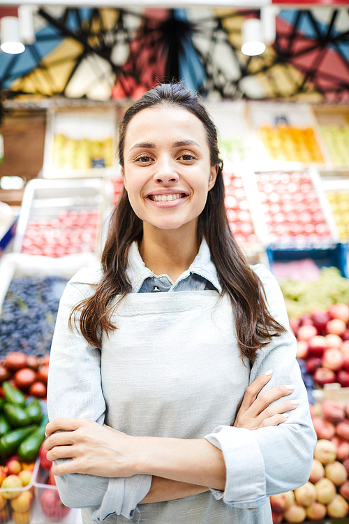 Positive confident young female grocer in apron crossing arms on chest and  while standing against fresh food stall at farmers market