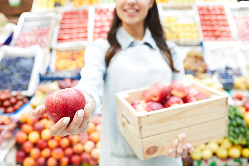Close-up of smiling female grocer in apron standing against organic food stall and holding wooden box full of apples, she giving fresh ripe apple to camera