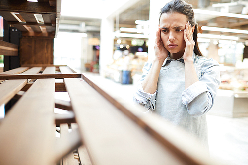 Sad frowning young woman massaging temples with fingers while feeling migraine and looking at empty shelf in farmers market