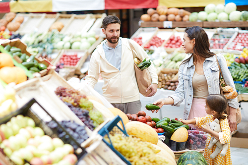 Young family buying fresh food at market: handsome bearded father asking wife while choosing vegetables, curious daughter listening to parents talk