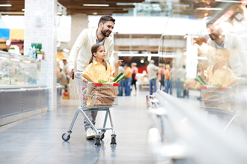 Serious cute girl sitting in shopping cart and pointing with finger at shelf while helping father with shopping in supermarket