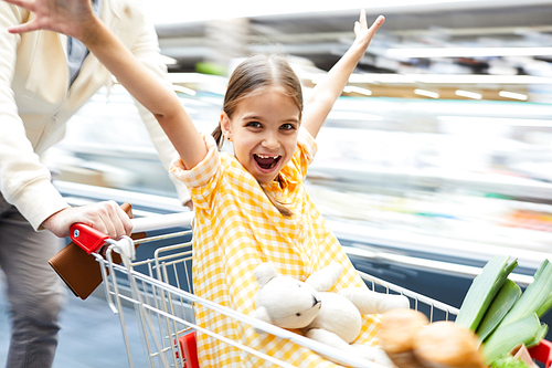 Jolly excited girl in checkered dress sitting in shopping cart and outstretching arms while having fun and  in supermarket, she riding in shopping cart