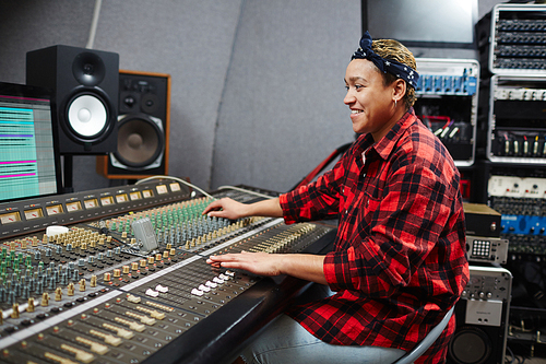 Smiling operator sitting by soundboard in studio, looking at monitor and mixing sounds