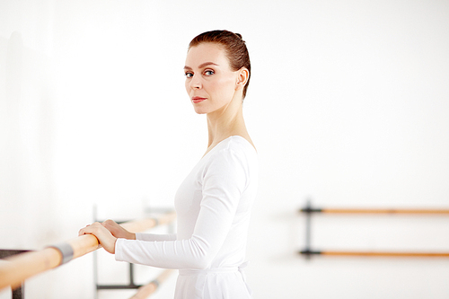 Beautiful ballerina in white clothes staying at wooden machine in dancing school and 