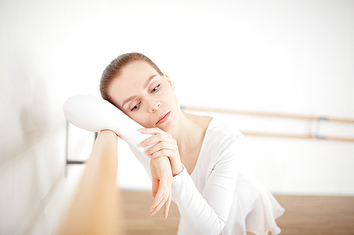 Adult ballerina staying at wooden machine with head on hands and thinking in light room