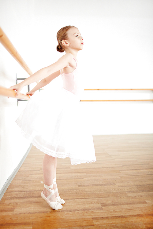 Little ballerina in fancy dress stretching her arms while dancing by bars