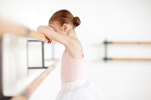 Tired little girl leaning against wooden bar in ballet classroom after training