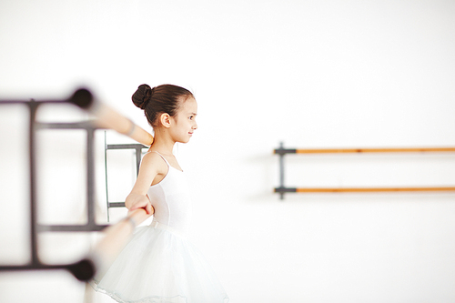 Side view young ballerina with strict hairstyle in white clothes smiling at wooden machine in dancing school