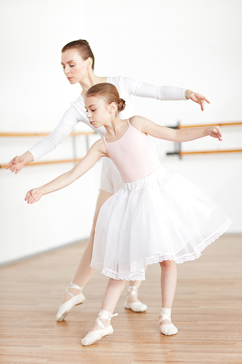 Woman and girl with strict hairstyle in light pointe and tutu staying in poses in white studio