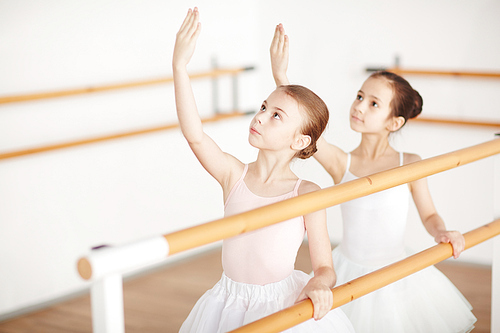 Small group of little girls in ballet dresses looking at their raised hands while training in classroom