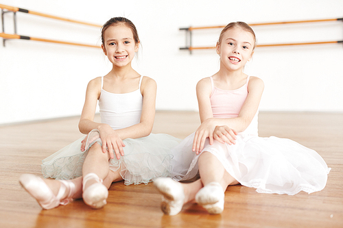 Adorable little ballet learners in white dressed sitting on the floor of classroom