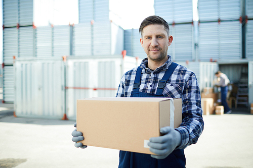 Content handsome manual worker in workwear holding cardboard box and  while working at container storage area