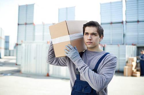 Serious handsome young mover in workwear carrying packaged product in box and holding it on shoulder while working at outdoor cargo warehouse