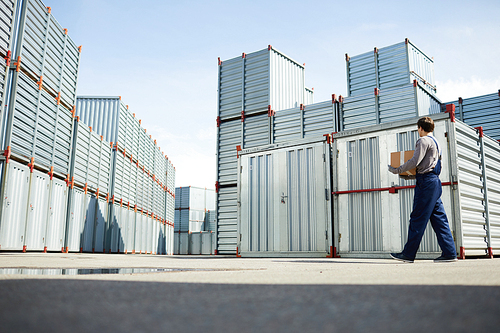 Young worker in workwear carrying big box while walking by several storage containers outdoors