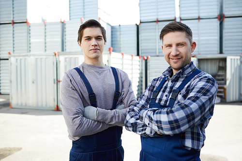 Two young delivery men with arms crossed on chest  on background of containers