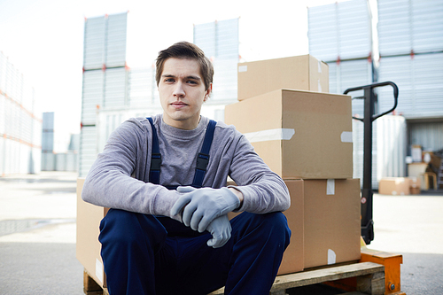 Serious handsome young male mover in workwear and gloves sitting on edge of load car and  in cargo container area