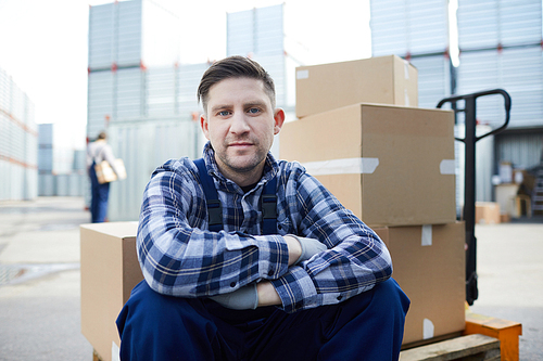 Restful loader man in workwear sitting on load cart with heap of packed carton boxes behind