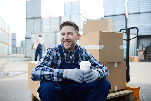 Cheerful excited young manual worker with stubble sitting on load cart and laughing while drinking coffee during break at outdoor storage container area