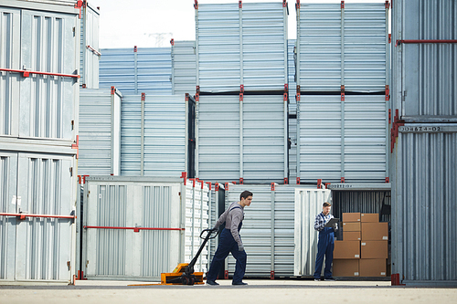 Young worker pulling empty load cart during loading work by metallic storage containers