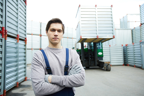 Cross-armed worker in grey pullover and dark blue overalls  on territory of transportation company
