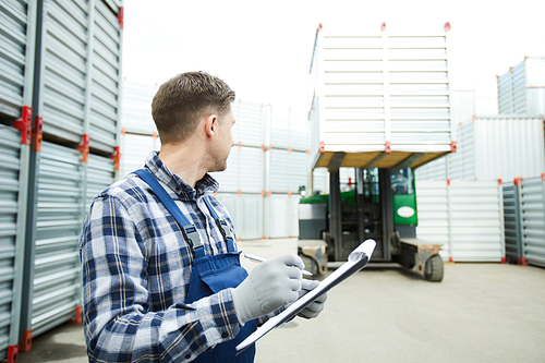 Contemporary worker in uniform making notes in document while seeing off machine with storage container