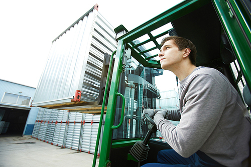 Serious focused handsome young man operating forklift while transporting heavy container over outdoor warehouse area
