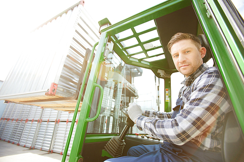 Serious handsome brutal forklift driver with stubble holding steering wheel and  while working at cargo area