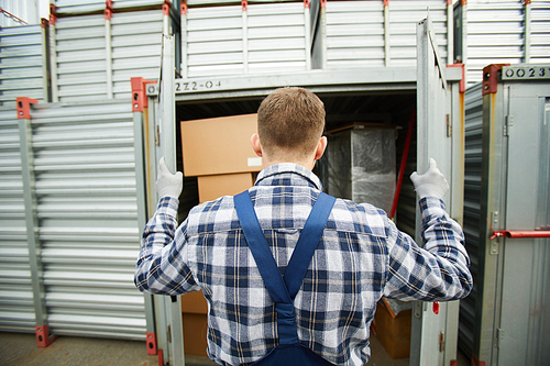 Rear view of busy man in checkered shirt opening metal cargo container and examining its content before shipment