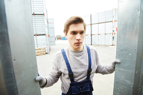 Frowning handsome young manual worker in workwear accepting cargo and opening container at shipment area
