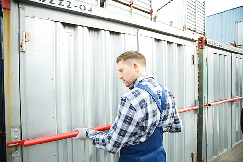 Worker in overalls checking safety bar while closing one of storage containers