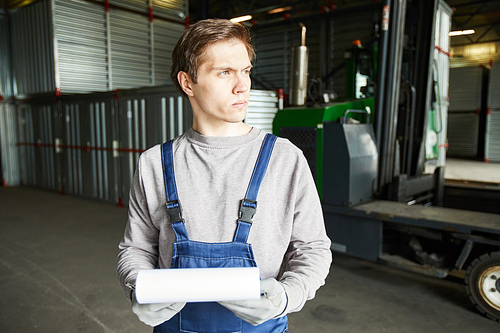 Serious pensive handsome male cargo storage worker in uniform holding clipboard and looking away while standing in storage room