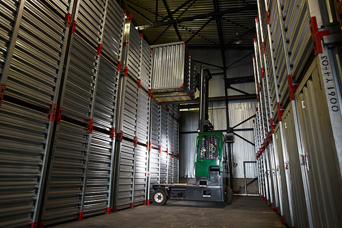 Green powerful forklift truck lowering large metal container in dark cargo storage area