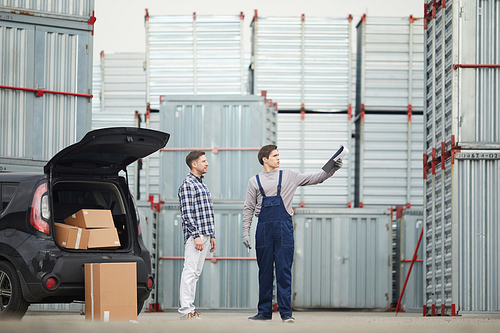 Outdoor warehouse manager in blue overalls holding clipboard with papers and pointing aside while explaining route to buyer standing at car with open trunk