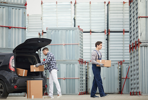 Serious young manual worker in overalls carrying boxes from car of client to container while man in white jeans taking boxes out of car trunk outdoors