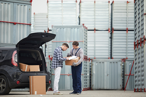 Concentrated young man in casual clothing signing contract with storage company after discussing deal: container storage worker holding box with clipboard and pointing where signature place is