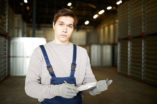 Serious handsome young male warehouse manager in workwear making notes in report and  while standing against cargo containers