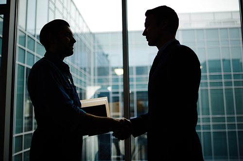 Silhouettes of businessman and delivery man with box shaking hands by office window
