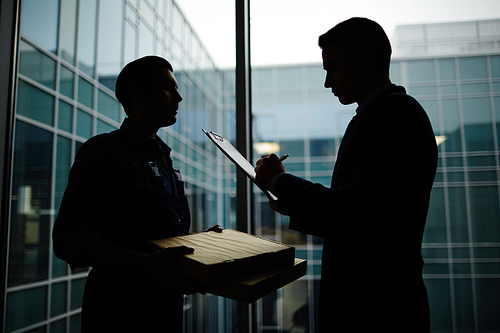 Contemporary businessman signing delivery paper by window with courier standing near by