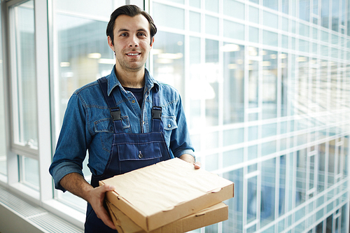 Contemporary young delivery service worker in uniform holding two packages with pizza for clients