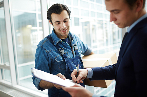 Young delivery service worker with box looking at businessman signing document of receiving package