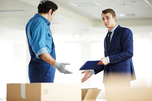 Young businesswoman with document pointing at open box while talking to relocation service staff in new office